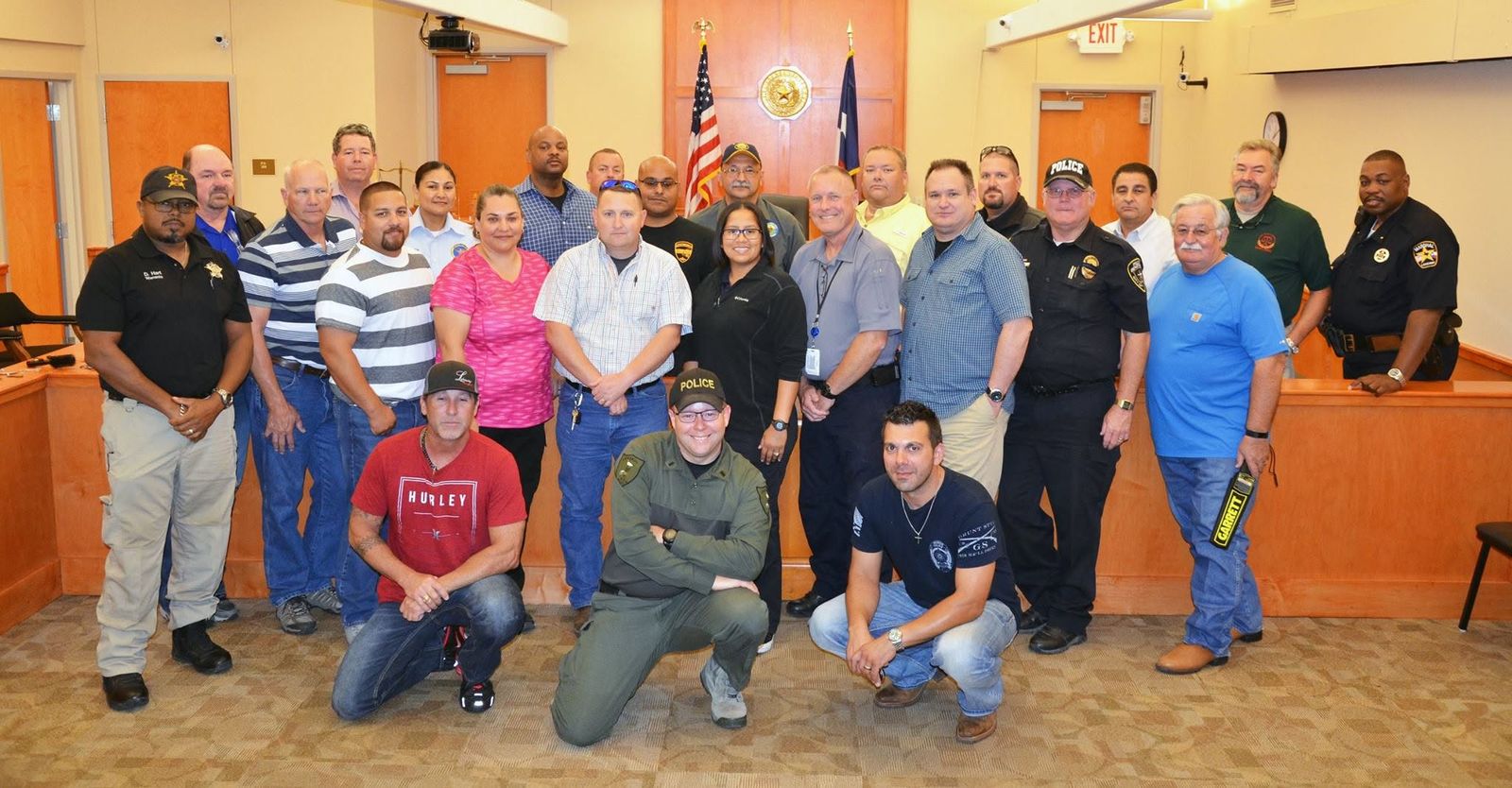A Court Security Specialist class assembled in a Texas courtroom at the end of the week. Roughly two dozen officers in uniform and plain clothes stand together before the bench, beneath the state seal and the United States and Texas flags.