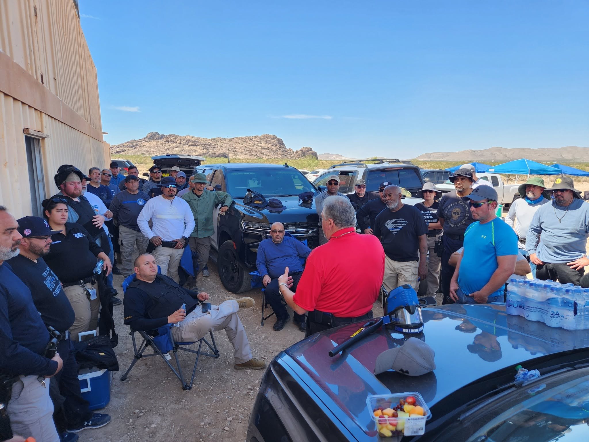 A class gathered around an instructor in the shade of a building for a debrief between scenarios.