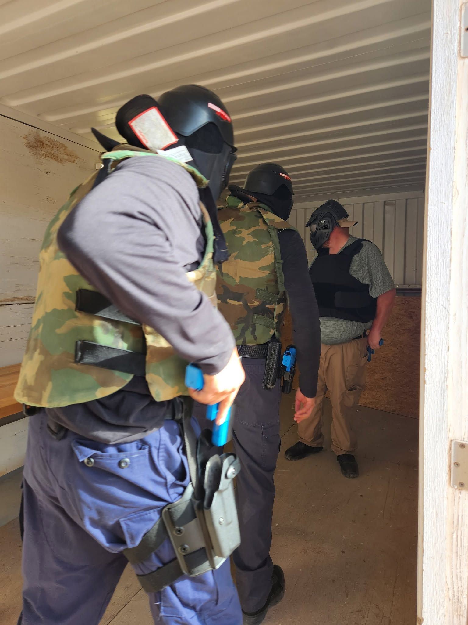 Three officers in protective masks and vests stacked at a doorway with marking-ammunition training weapons.