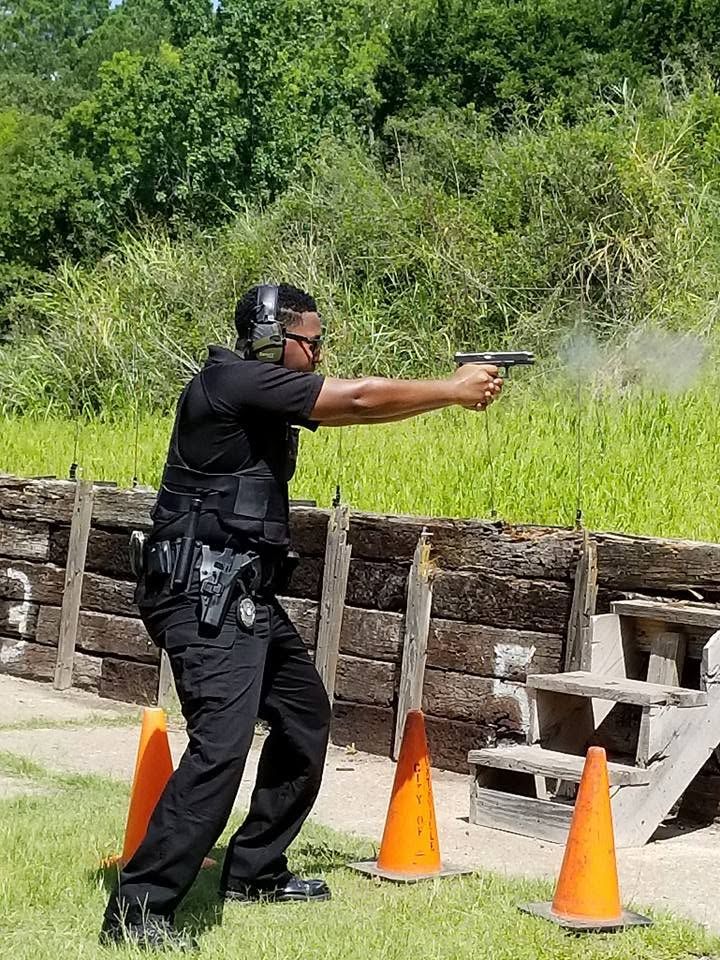 A court security officer in protective headgear and a chest rig firing a handgun on the range during the live-fire practical, smoke still at the muzzle.